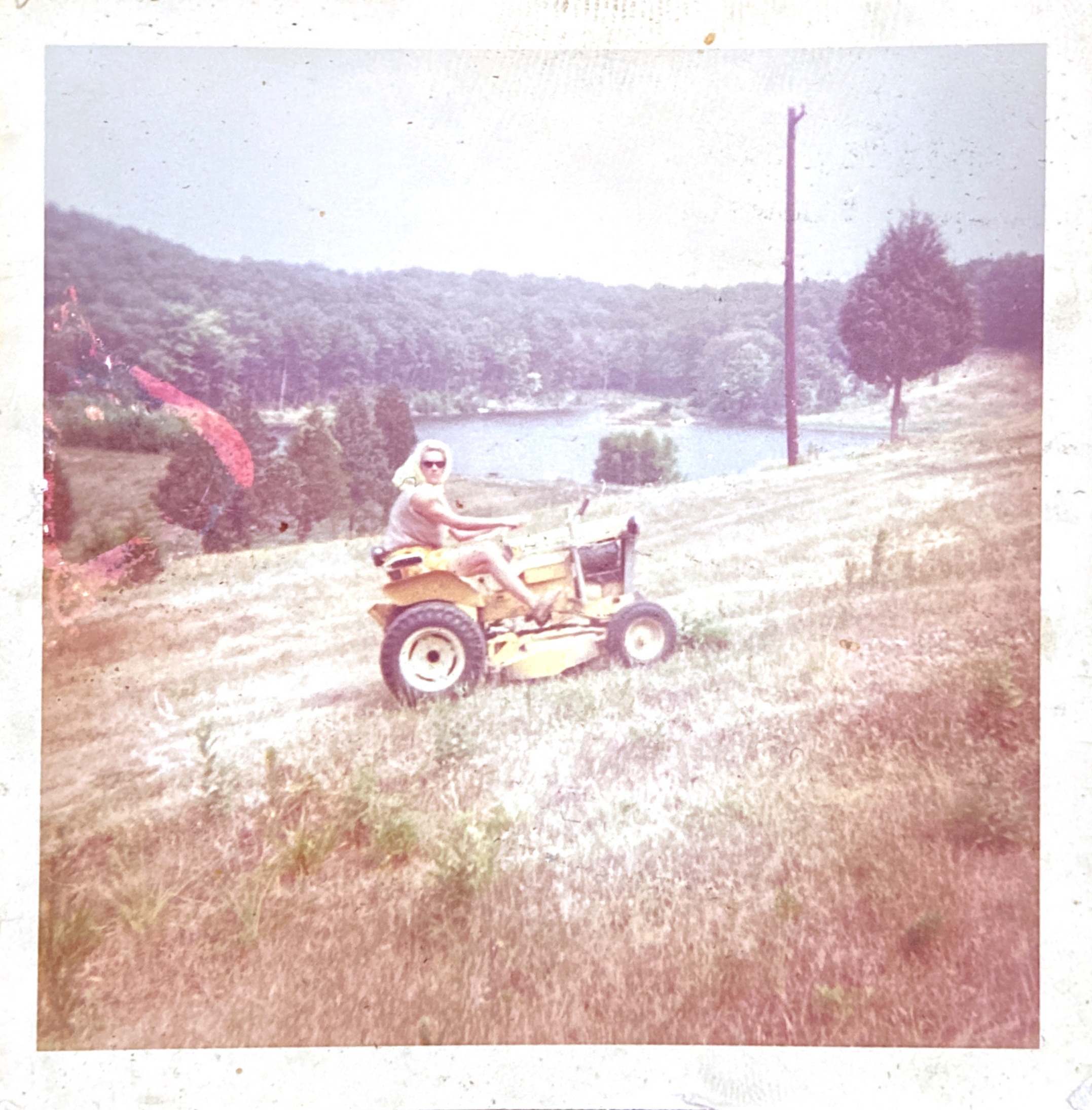 Summer 1966, Barbara Beans (Bob’s wife) mowing the hillside between their loghouse and the lake. That’s our B-10 tractor, which Tom did most of his mowing with.